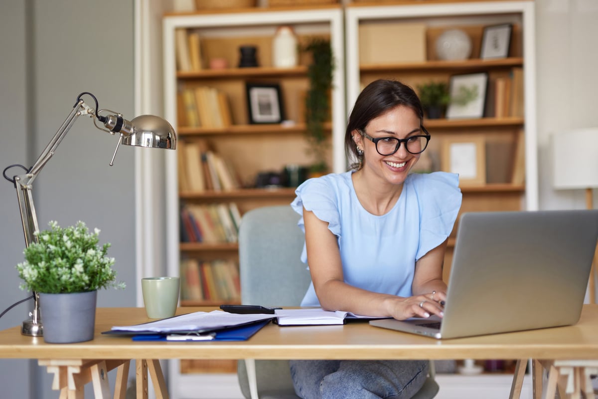 Smiling young woman working from home, typing on a laptop at a cozy desk